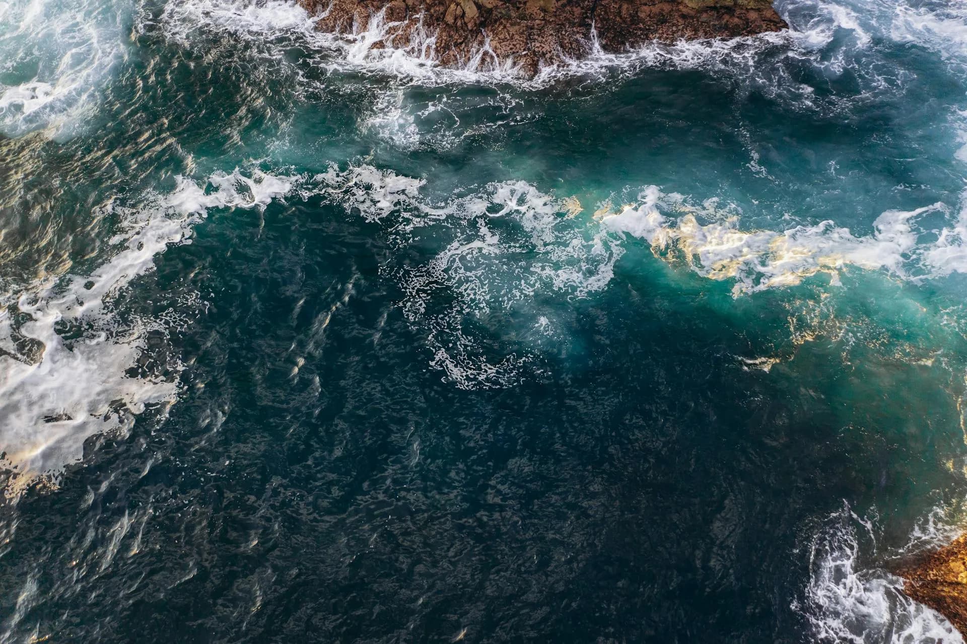 Dramatic aerial shot of ocean waves crashing against rocky shoreline, capturing the vibrant blue and white waters.